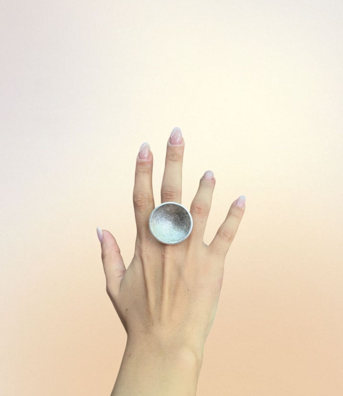 A hand wearing the Lunar Shell Ring — a large, concave, silver-plated ring with a soft textured interior, shaped like a moon crater or seashell.