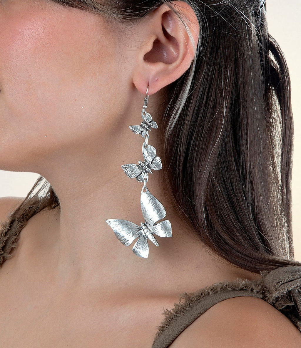  Close-up of a silver earring featuring three detailed butterfly charms, worn on a woman's ear with dark hair and a soft top.