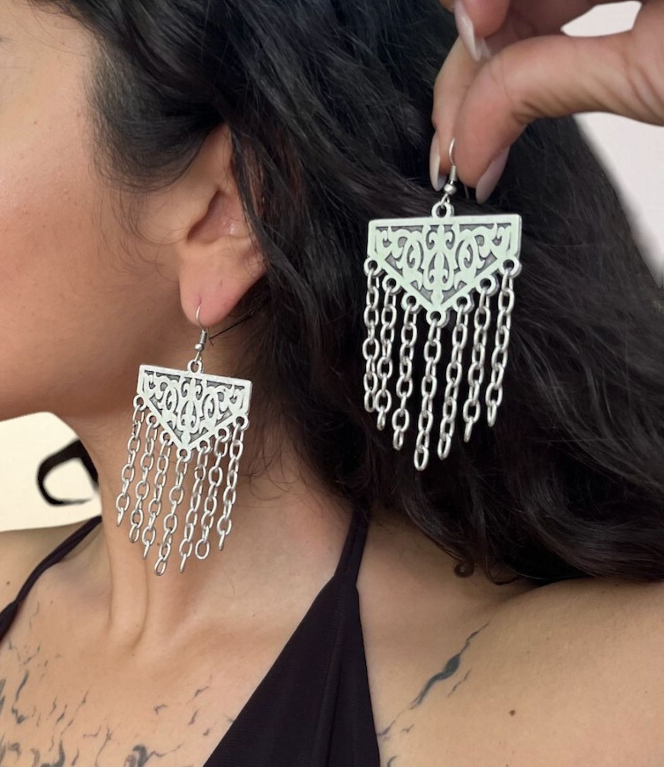 A close-up of a woman wearing a large silver earring featuring an ornate triangular top and cascading chain fringe, with another matching earring held beside her face.