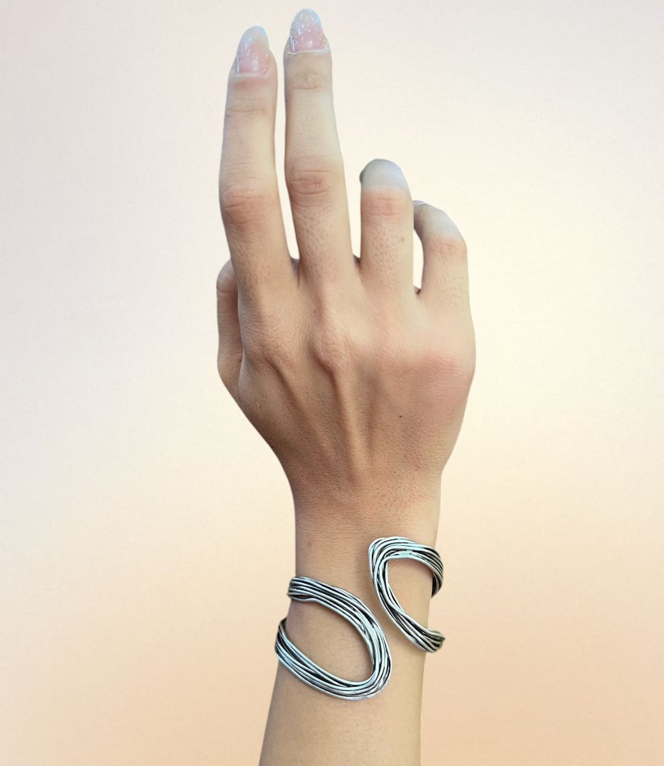 A silver-tone open cuff bracelet with sculptural, flowing wave shapes worn on a wrist against a soft gradient background.