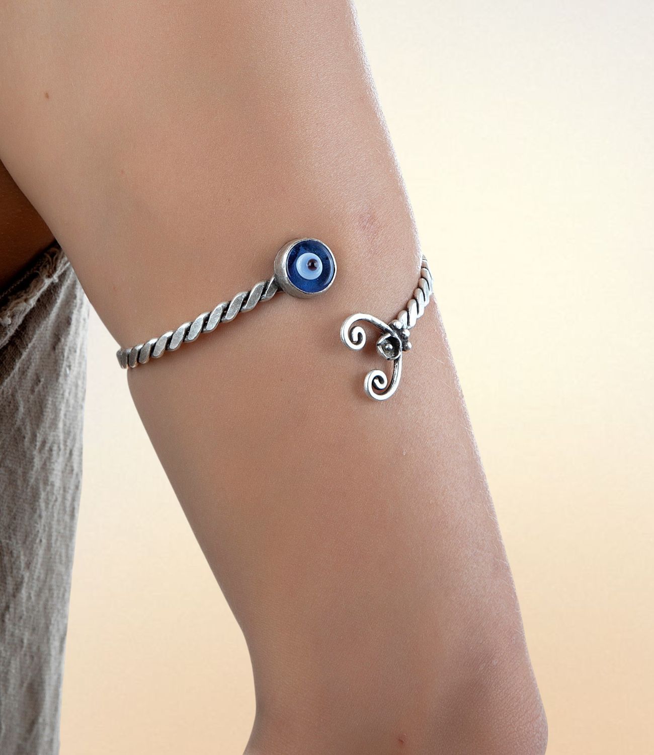Silver twisted arm cuff featuring a central blue evil eye bead and decorative spirals, worn on the upper arm.
