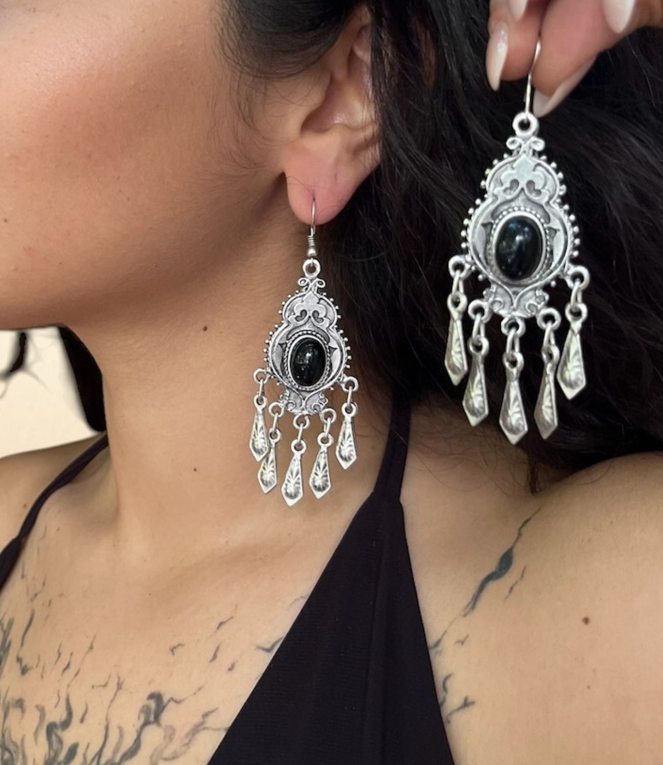 Ornate silver chandelier earring with a black onyx center and five hanging silver leaf-shaped charms, worn by a woman with dark hair. A matching earring is held close to the ear for emphasis.
