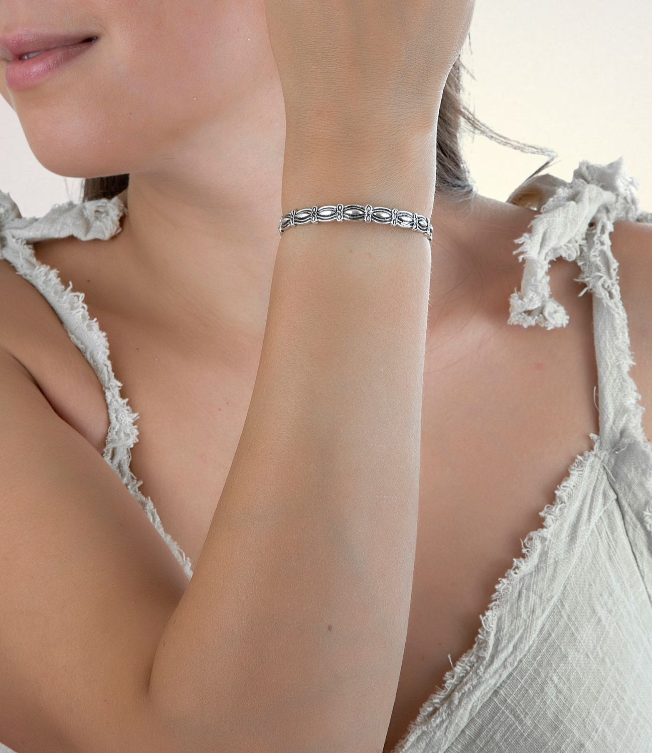  close-up of a slim silver bracelet with detailed oval links on a woman’s wrist, styled against a soft white sleeveless top.