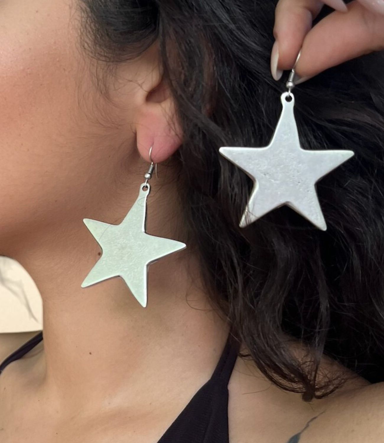 a person wearing large silver star-shaped earrings, one earring shown clearly in hand to highlight its bold size and shiny texture.