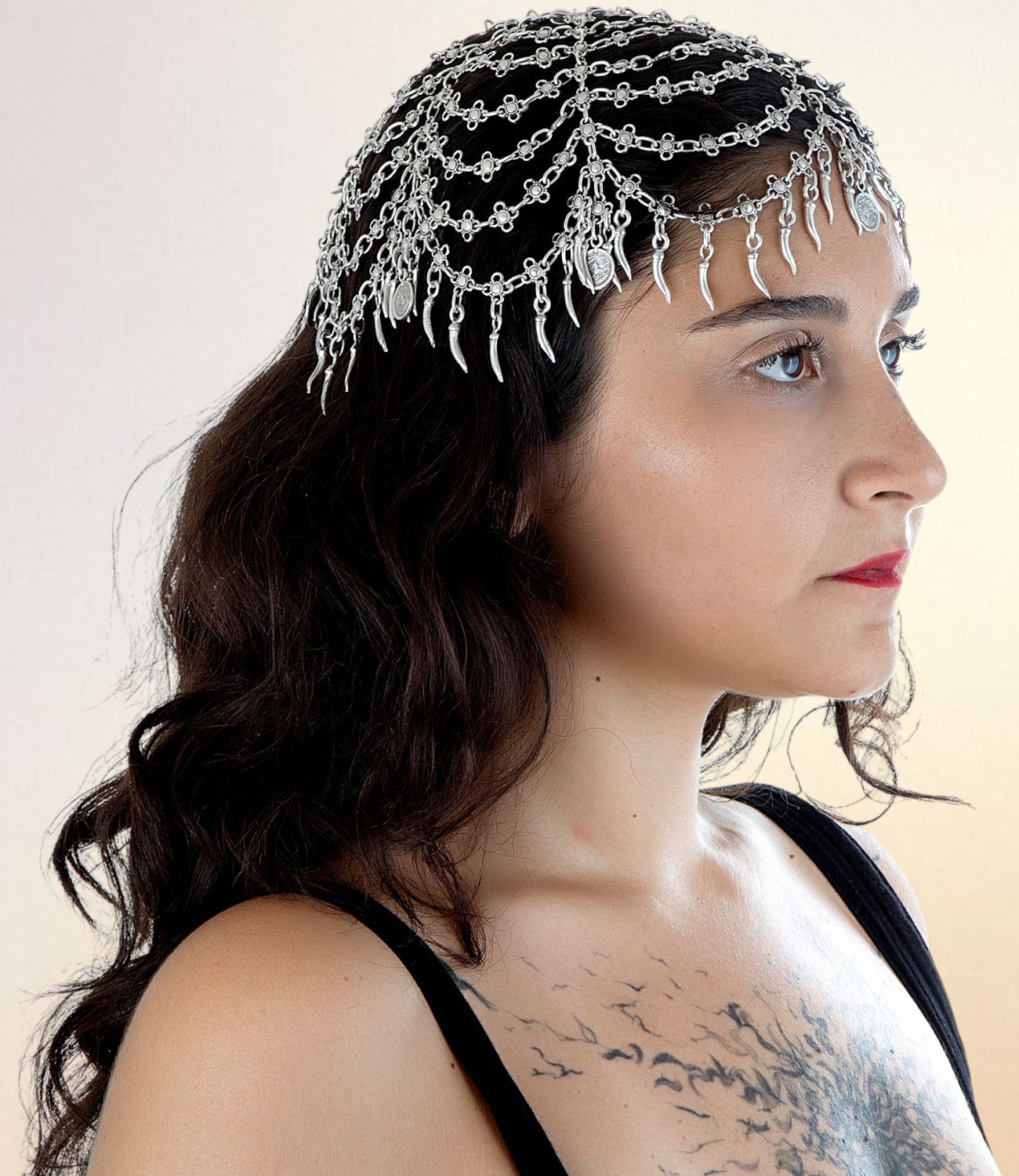 Side profile of a woman wearing a silver head chain with layered rows of links and hanging charms across her forehead and hair, styled against a neutral background.