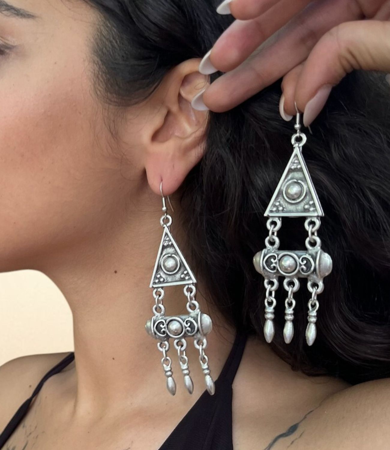  A silver statement earring featuring a triangle top, ornate cylinder center, and three pointed drops hanging below, worn by a woman with dark hair. A second matching earring is held near the face for display.