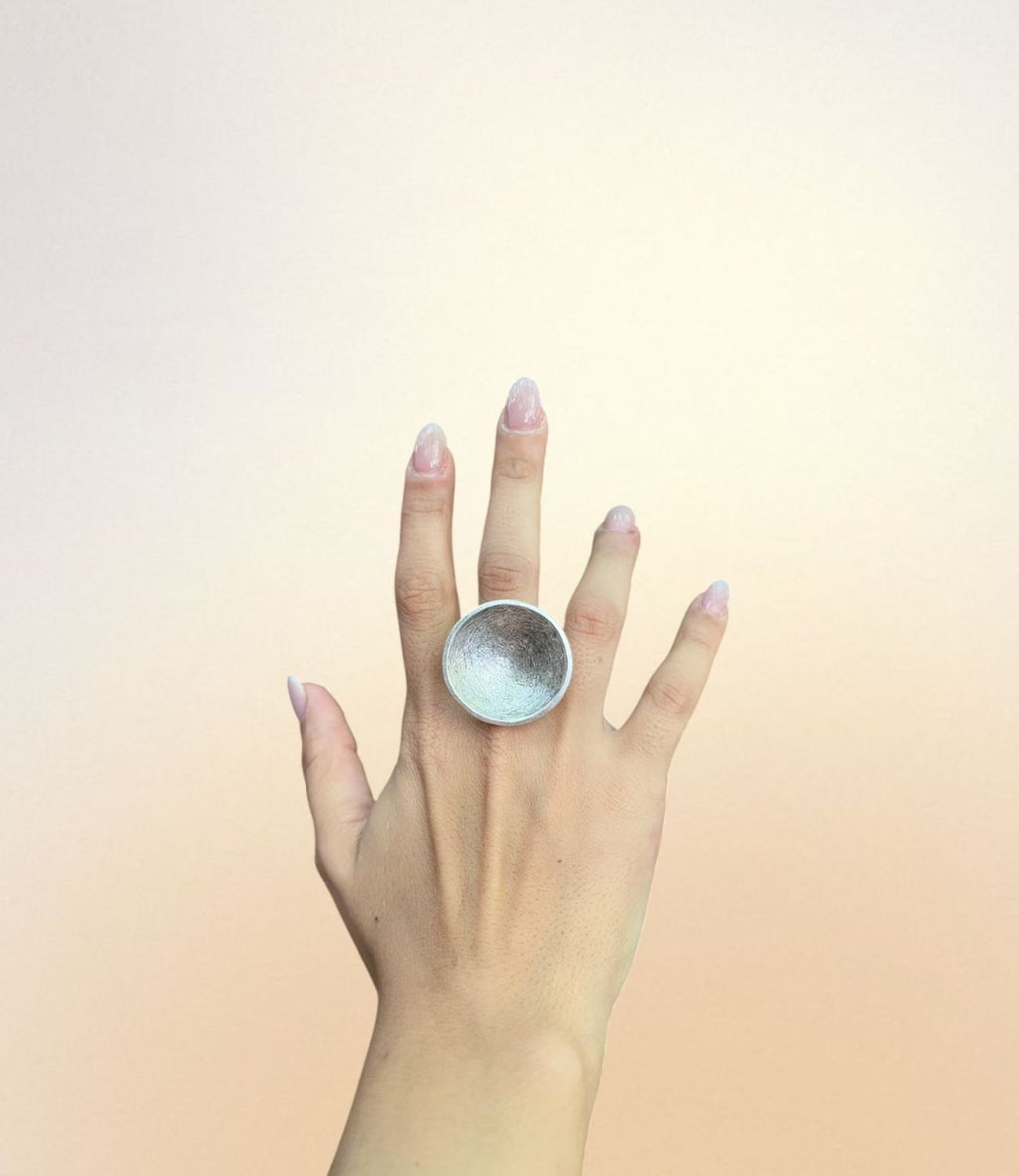 A hand wearing the Lunar Shell Ring — a large, concave, silver-plated ring with a soft textured interior, shaped like a moon crater or seashell.