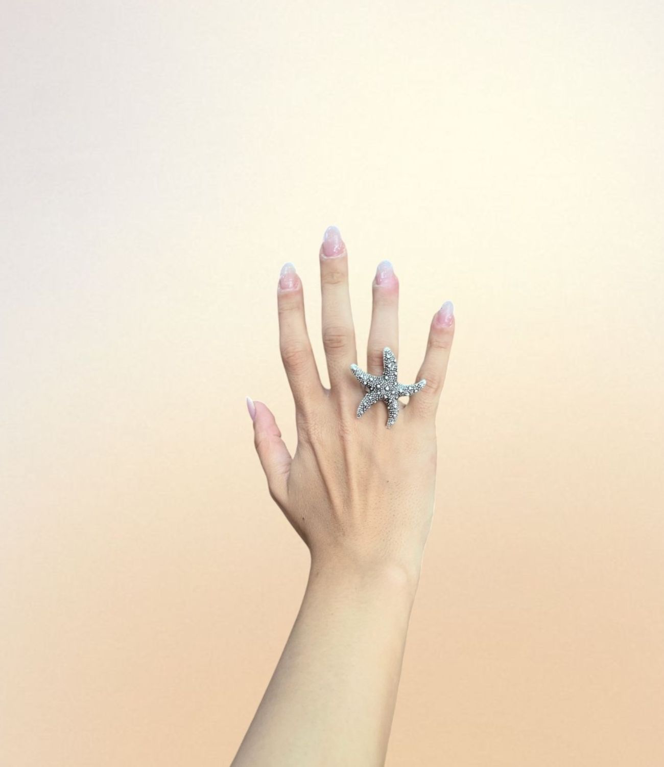 A hand wearing the Starfish Ring — a large, silver-plated ring in the shape of a textured starfish, covering two fingers and evoking beachy, oceanic vibes.